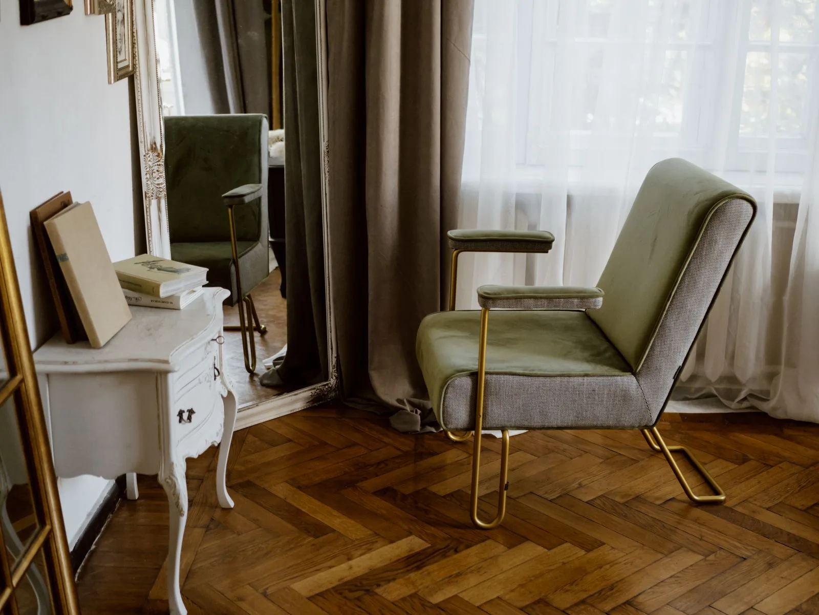 A contemporary green velvet and brass armchair beside an ornate white French antique console and gilt mirror on a herringbone parquet floor