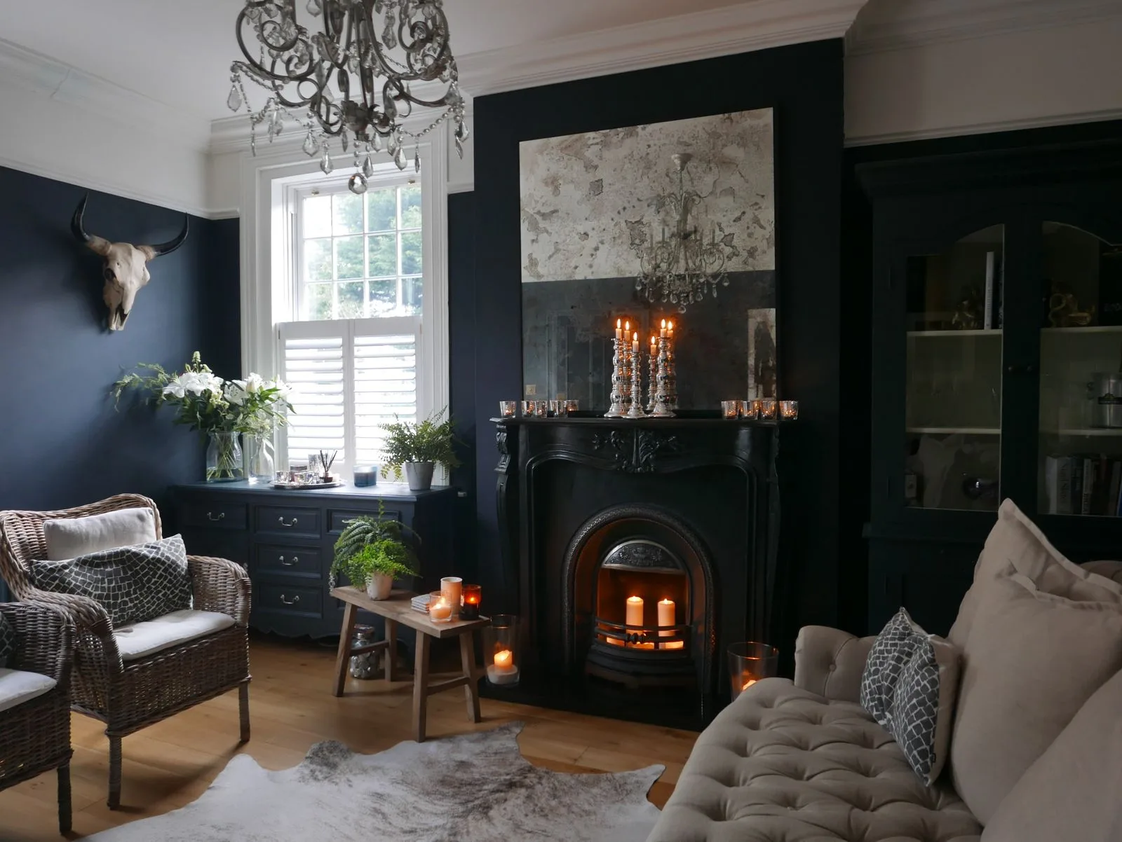 A navy sitting room mixing an antique black fireplace, aged mirror and chandelier with a contemporary buttoned sofa